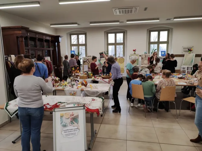Warsztaty rękodzieła - na zdjęciu, po lewej stronie, wystawa prac rękodzielnicznych (na stołach), a po prawej stronie dzieci wraz z osobami dorosłymi tworzące kolejne prace rękodzielnicze
