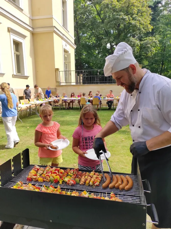 Na zdjęciu kucharz w trakcie nakładania kiełbasy z grilla na tackę trzymaną przez dziewczynkę